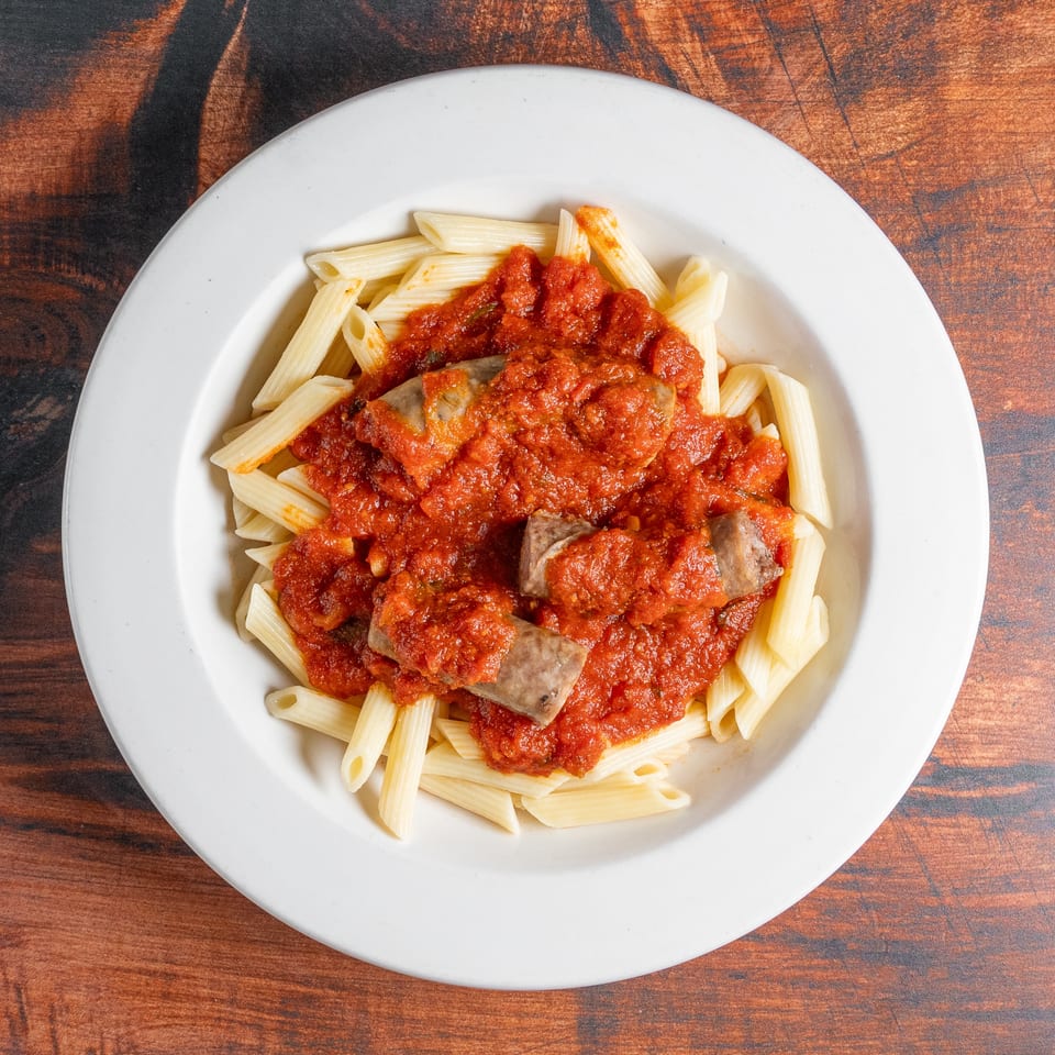 Penne pasta with tomato sauce and sausage pieces in a white bowl on a wooden table.