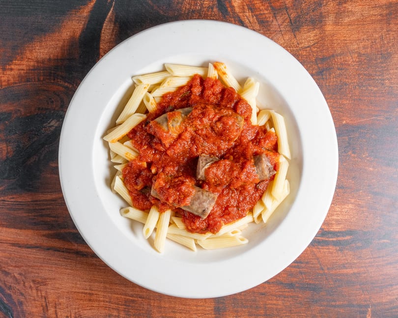 Penne pasta with tomato sauce and sausage pieces in a white bowl on a wooden table.
