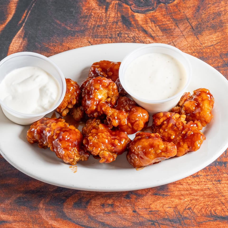 A white plate holds boneless wings coated in a glossy orange sauce, with two cups of ranch dressing.