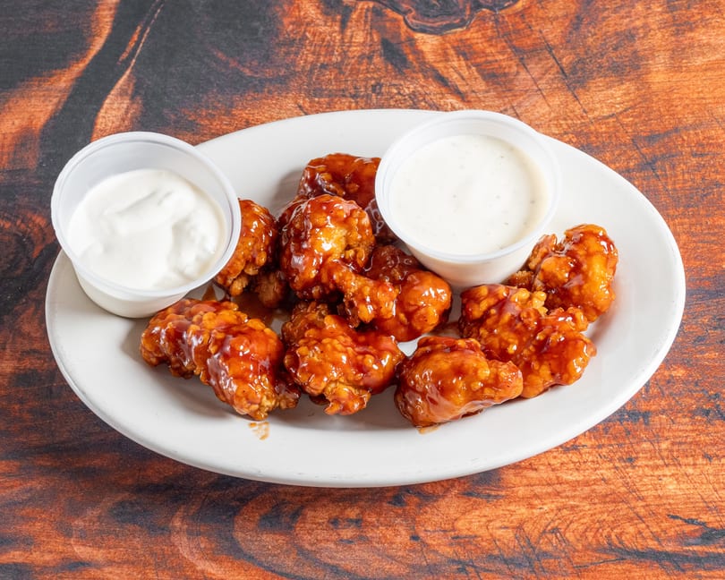 A white plate holds boneless wings coated in a glossy orange sauce, with two cups of ranch dressing.