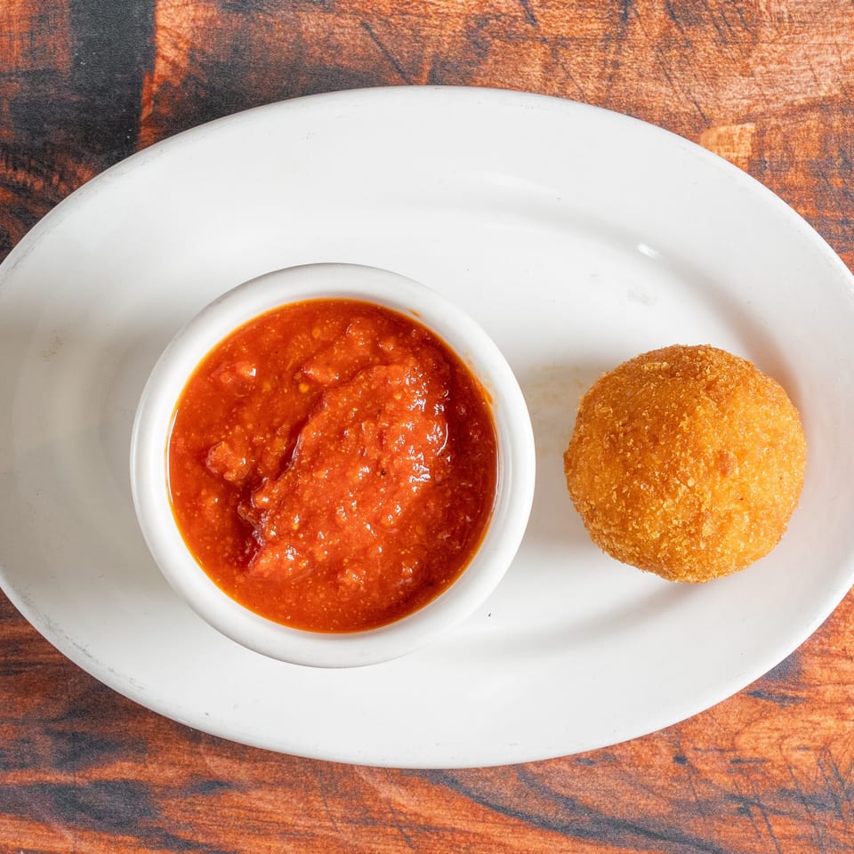 Golden-brown arancini ball and a white ramekin of red marinara sauce on a white plate.