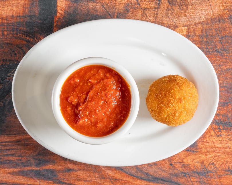 Golden-brown arancini ball and a white ramekin of red marinara sauce on a white plate.