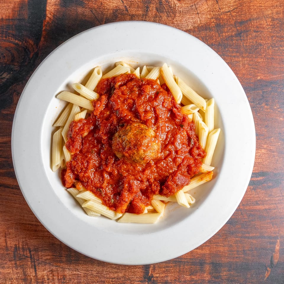 Penne pasta with rich red marinara sauce and a single meatball in a white bowl.