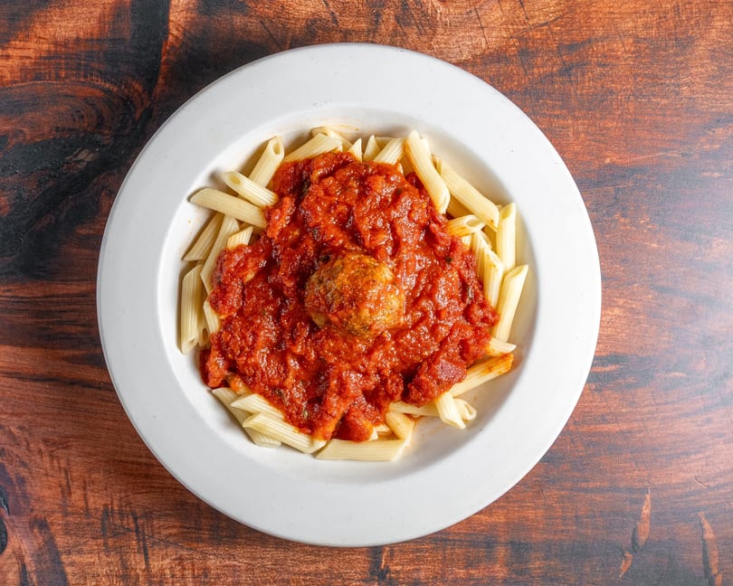 Penne pasta with rich red marinara sauce and a single meatball in a white bowl.