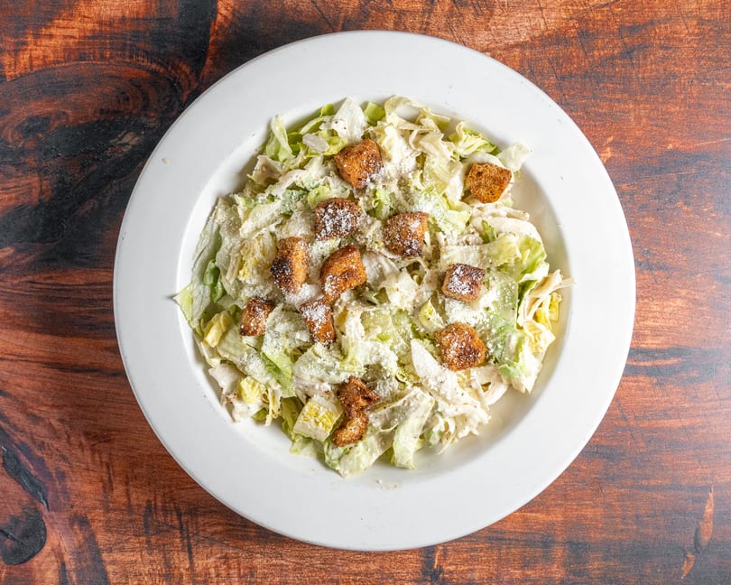 Overhead view of a Caesar salad with lettuce, croutons, and parmesan cheese in a white bowl.