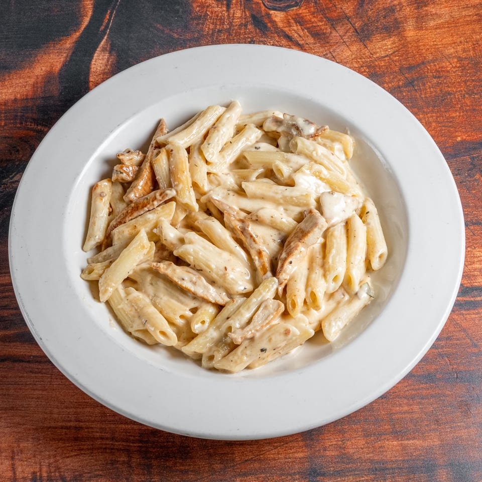 Creamy penne pasta with chicken pieces in a white bowl on a dark wooden table.