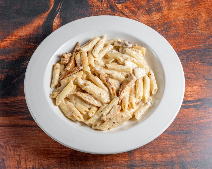 Creamy penne pasta with chicken pieces in a white bowl on a wooden table.