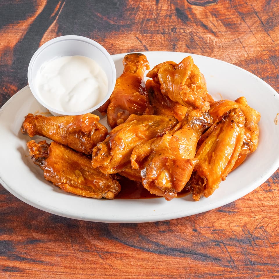 A white plate holds several saucy chicken wings and a small cup of white dipping sauce.