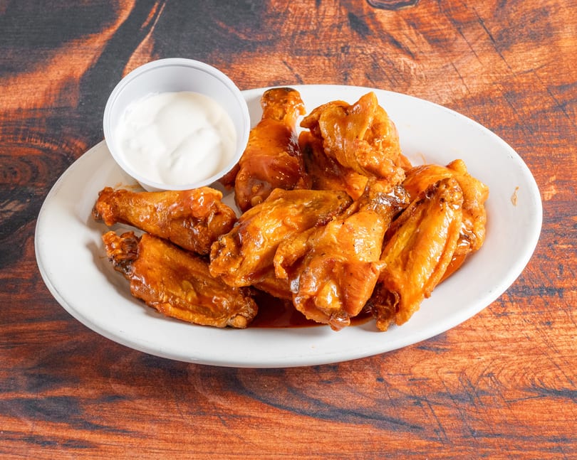 A white plate holds several saucy chicken wings and a small cup of white dipping sauce.