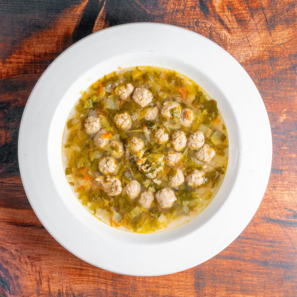 A white bowl of Italian wedding soup with meatballs and green vegetables on a wooden table.