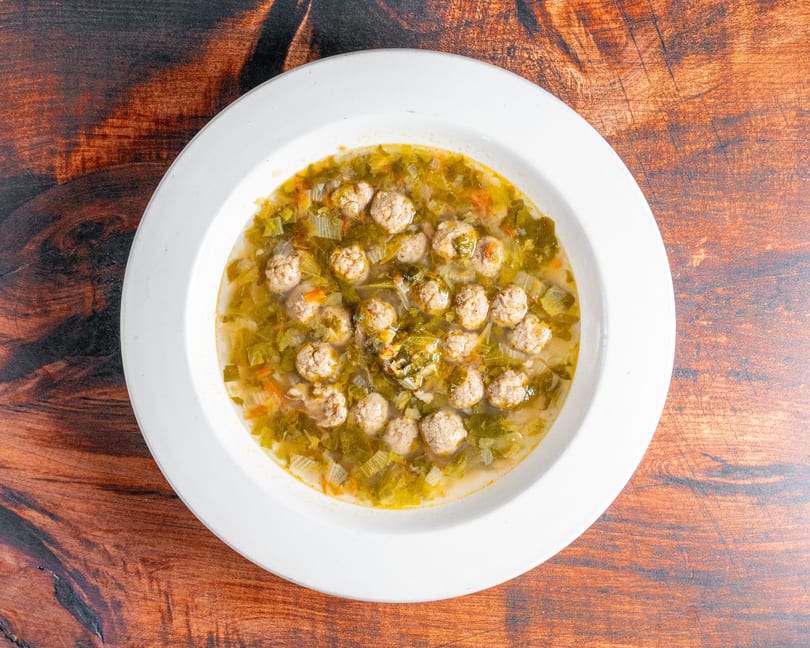 A white bowl of Italian wedding soup with meatballs and green vegetables on a wooden table.
